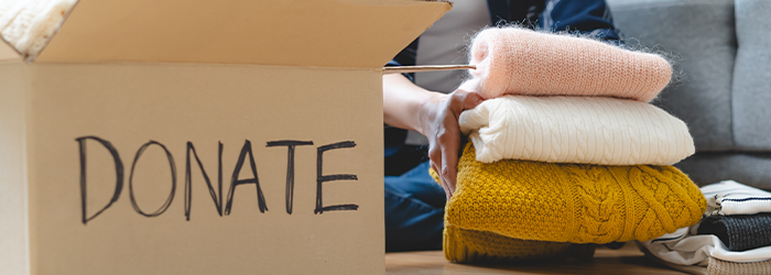 Cardboard box with word Donate on it and woman holding stack of three different color folded sweaters