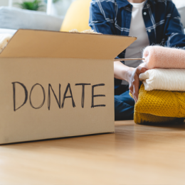 Cardboard box with word Donate on it and woman holding stack of three different color folded sweaters